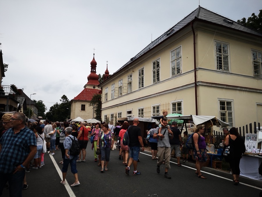 Budova školy v létě, před ní ulice plná lidí při trhu, v pozadí věž kostela